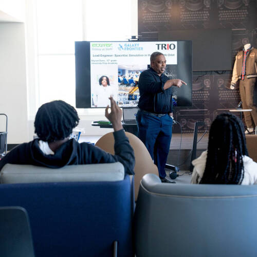 A presenter gestures toward a slide on space engineering while students in a lounge area listen, one raising a hand to ask a question.